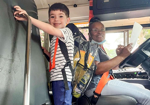 Young child wearing clear backpack getting on the school bus while driver watches.