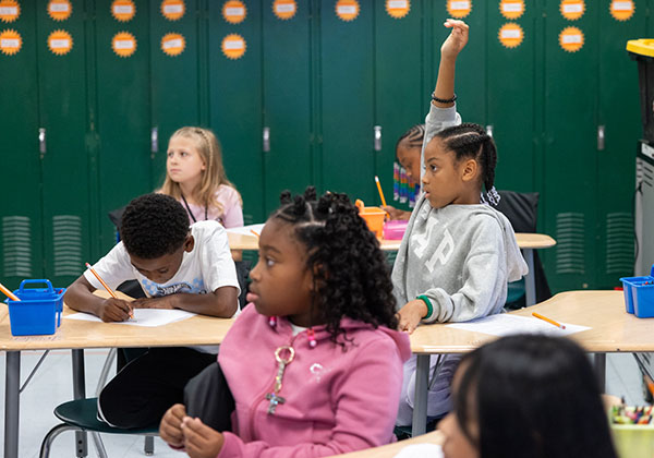 A student raises her hand in a classroom with other children.