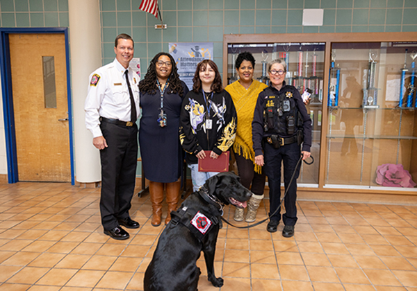 Two school staff members, a student and law enforcement officers pose together in a school lobby with a black Labrador service/working dog wearing a vest.